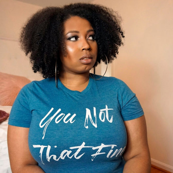 An African American woman in a pale pink bedroom with a medium sized curly afro looking off to the right with a sarcastic expression. 